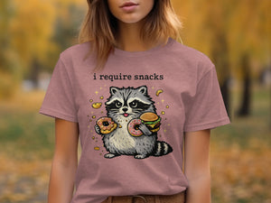 T-shirt graphic featuring a raccoon holding donuts and a burger with "I require snacks" text.