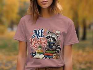 Cute raccoon "All Booked" graphic with books, coffee, and glasses on a pink t-shirt.
