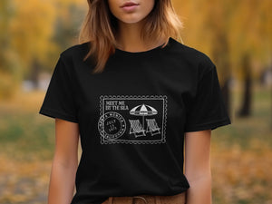 Black t-shirt with "Meet Me By The Sea" stamp graphic with beach chairs and umbrella.