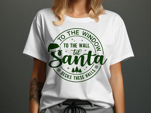 White t-shirt with green text "To the window to the wall til Santa decks these halls".