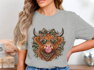 Highland cow wearing heart sunglasses and blowing bubblegum on a grey t-shirt.
