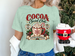 Light green t-shirt graphic with "Cocoa Social Club" and hot cocoa mug.