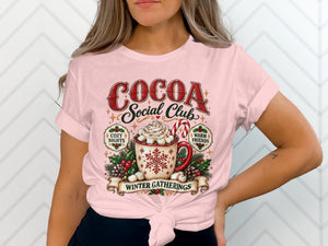 Pink t-shirt with "Cocoa Social Club Winter Gatherings" graphic and festive mug.
