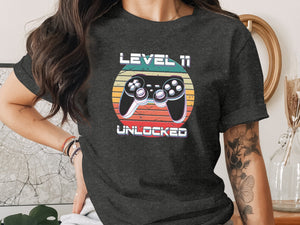 "Level 11 Unlocked" text with retro joystick and sunset background on a dark t-shirt.
