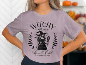 Light purple t-shirt with black graphic of a witch, bats, and text "Witchy Woman Social Club".