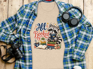 T-shirt with "All Booked" text and raccoon on books, laid flat with plaid shirt.
