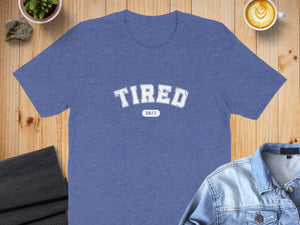 Blue t-shirt with "TIRED 24/7" graphic on a wooden surface.