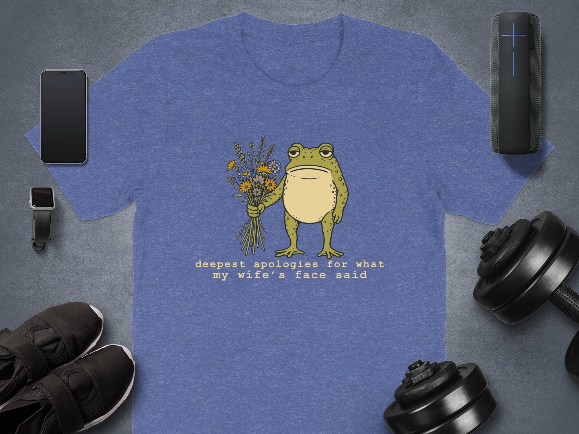 Blue t-shirt with drawing of frog holding flowers and text saying "deepest apologies for what my wife's face said".