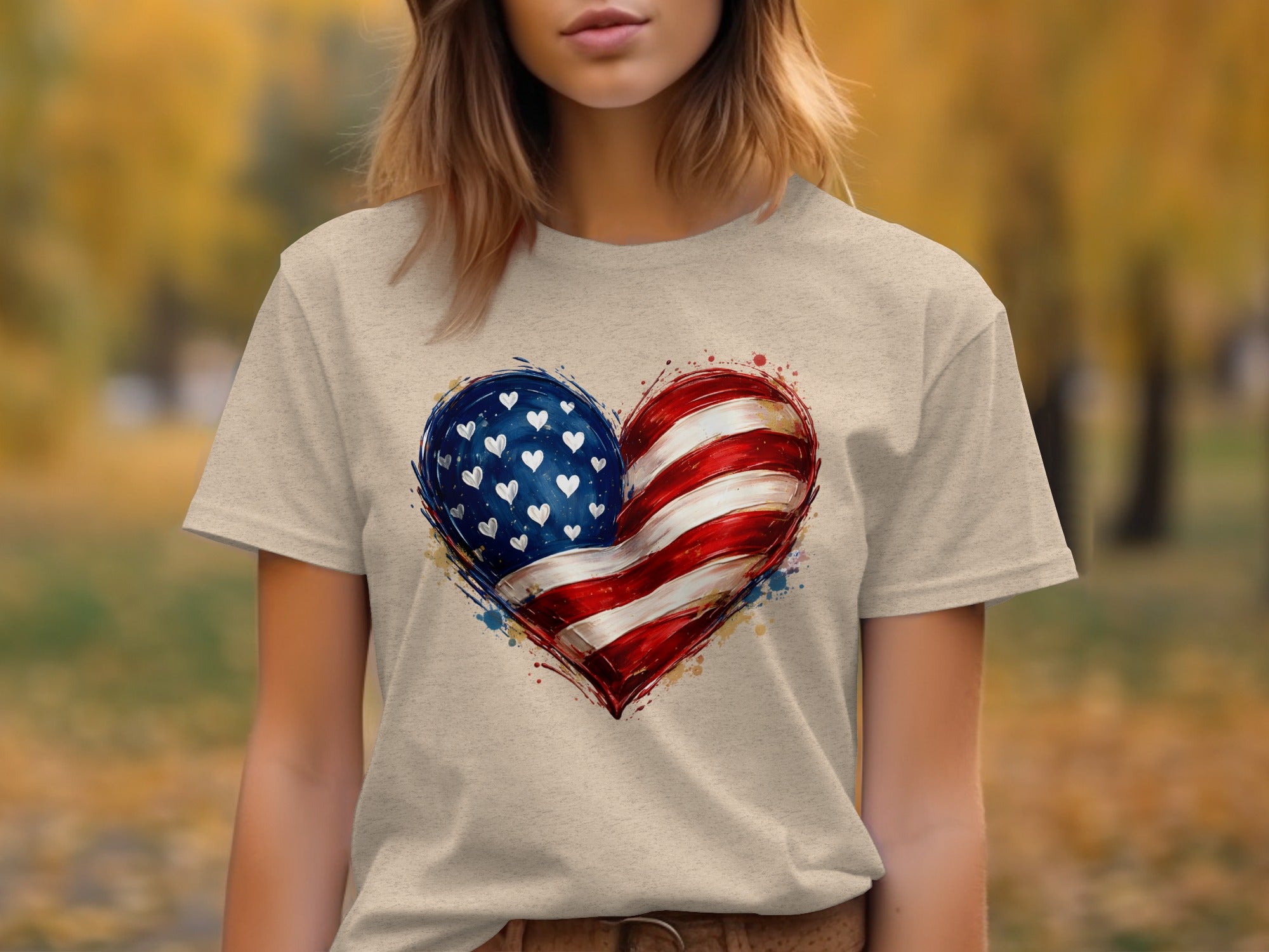 Light tan t-shirt with American flag heart graphic and a blurred autumn background.