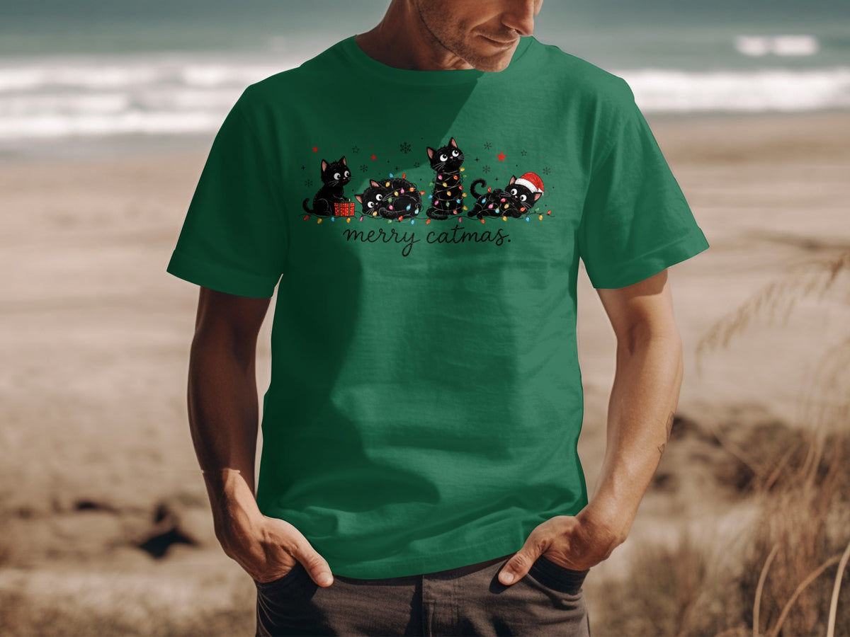 A man wearing a green T-shirt with a graphic of four black cats tangled in Christmas lights and the text merry catmas.
