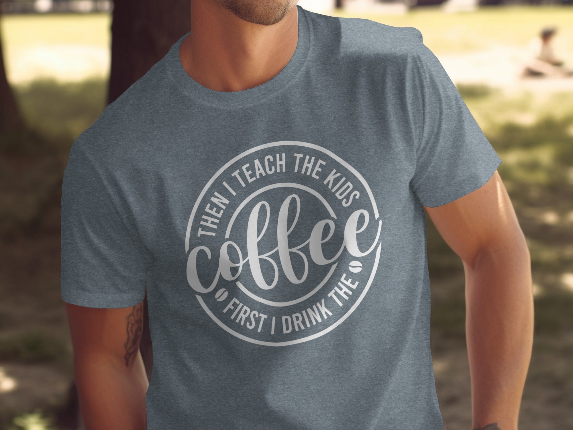 Grey t-shirt with a circular design that says "Coffee First I Drink The Then I Teach The Kids".