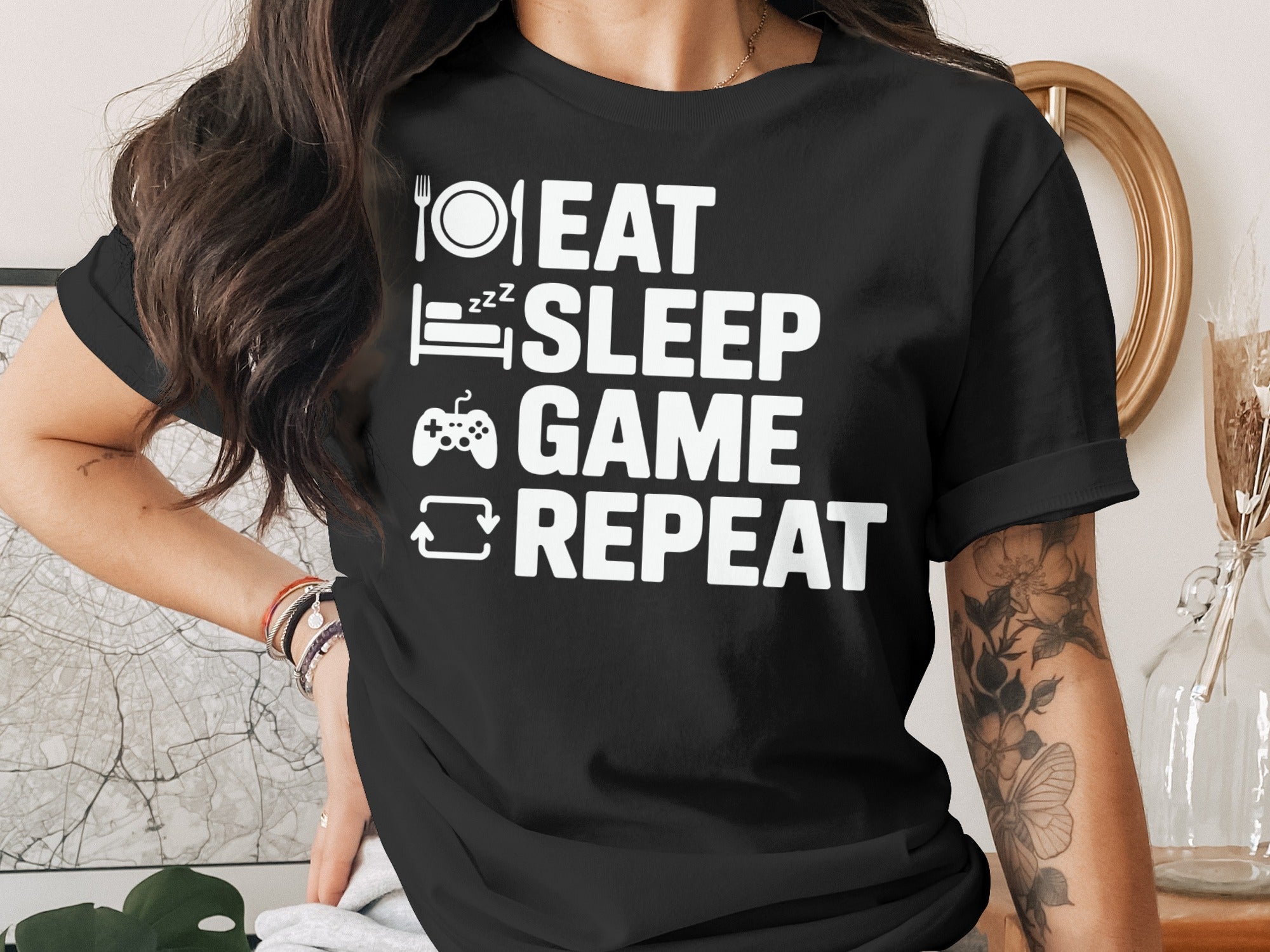 Black t-shirt with white text "Eat Sleep Game Repeat" and icons of a plate, bed, controller, and refresh symbol.