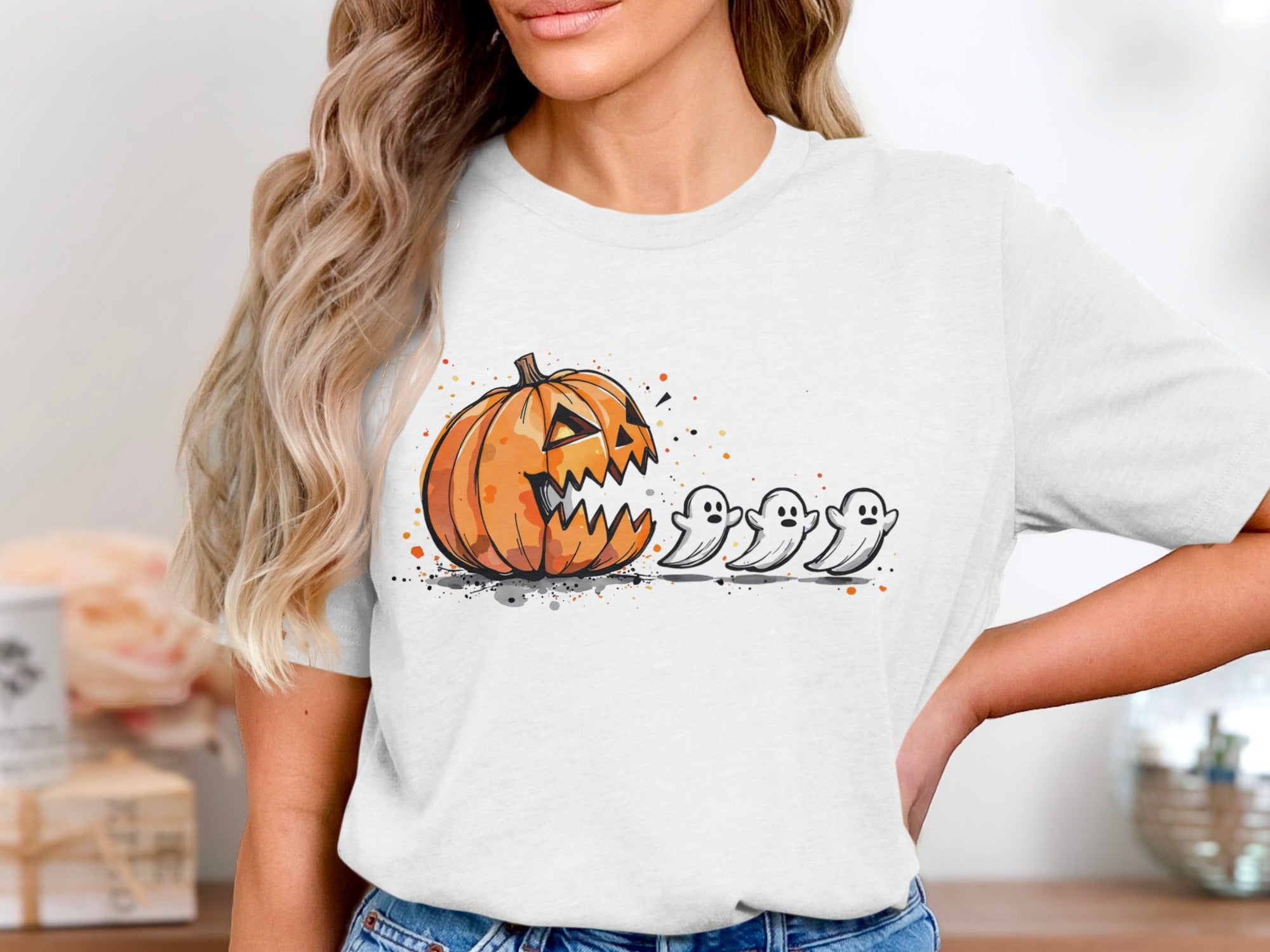 White t-shirt with a cartoon pumpkin chasing three ghosts with splatter paint accents.