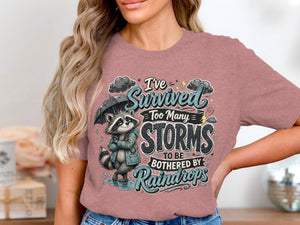 T-shirt graphic with raccoon under umbrella and text "I've survived too many storms to be bothered by raindrops".