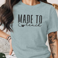 Light blue t-shirt with black text reading "MADE TO teach" and a small heart.