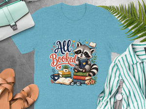 Light blue t-shirt with a watercolor graphic of a raccoon reading books, text "All Booked".