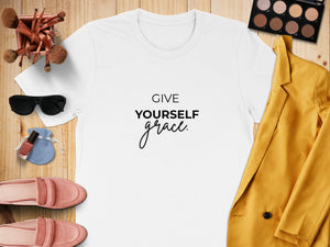 White t-shirt with "GIVE YOURSELF grace." text, flat lay with makeup and accessories.