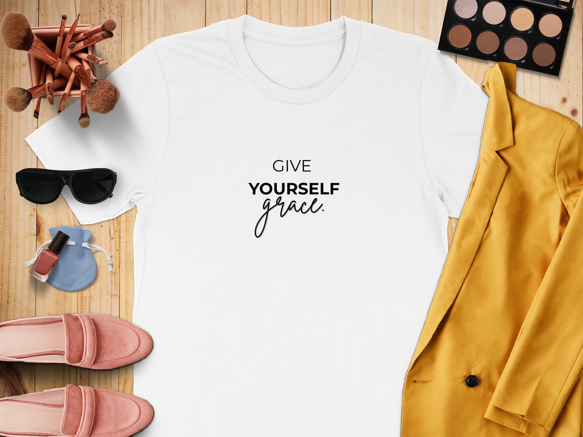 White t-shirt with "GIVE YOURSELF grace." text, makeup brushes, sunglasses, nail polish, and pink loafers.