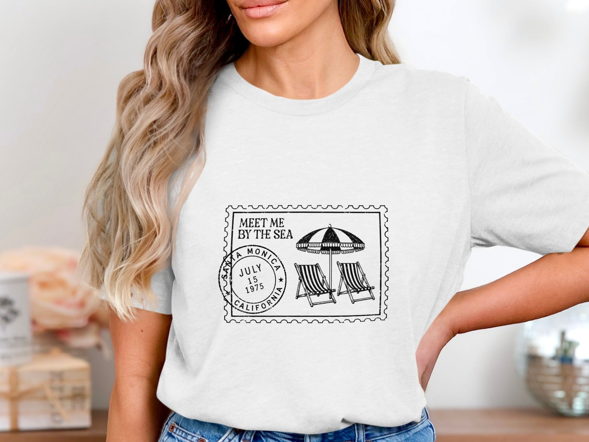 White t-shirt with stamp design: "Meet Me By The Sea," Santa Monica, July 15, 1975, California, beach umbrellas, and chairs.