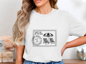 White t-shirt with stamp design: "Meet Me By The Sea," Santa Monica, July 15, 1975, California, beach umbrellas, and chairs.