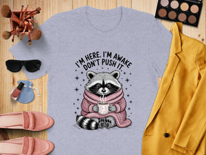 Grey t-shirt with illustration of raccoon holding coffee and text "I'm here, I'm awake".