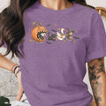 Halloween graphic with grinning pumpkin, ghosts, black cat, witch, and duck on purple shirt.
