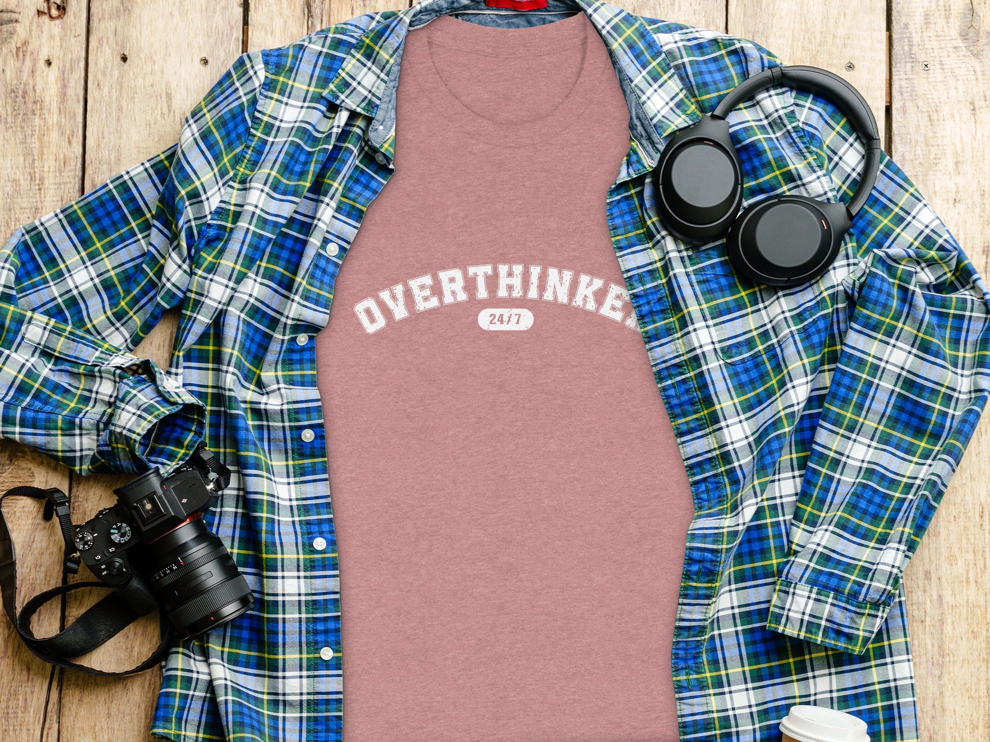 Pink t-shirt with "OVERTHINKER 24/7" printed, next to a plaid shirt, headphones, and camera.