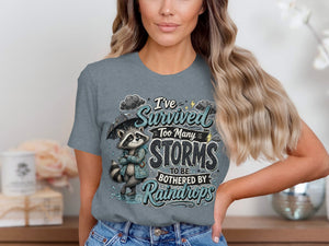T-shirt graphic with raccoon holding umbrella and text "I've survived too many storms to be bothered by raindrops".