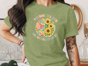Green t-shirt with a brain graphic split between colorful patchwork and sunflowers, text reads "The sun will rise and we will try again."