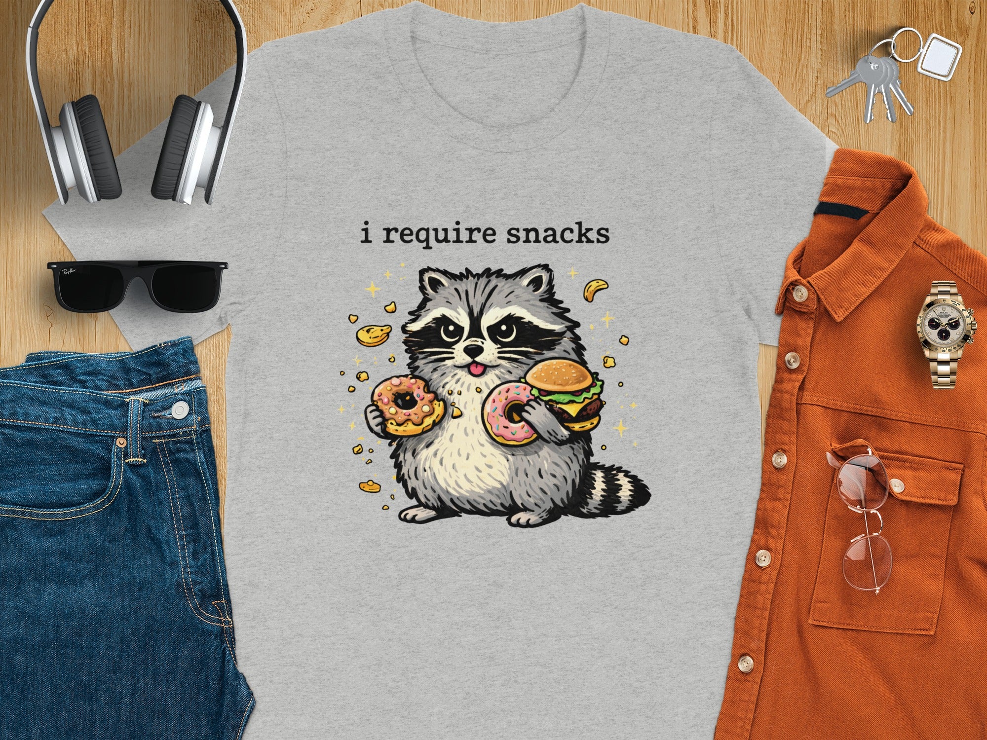 Grey t-shirt with a cartoon raccoon holding a burger and two donuts. Text reads "i require snacks."