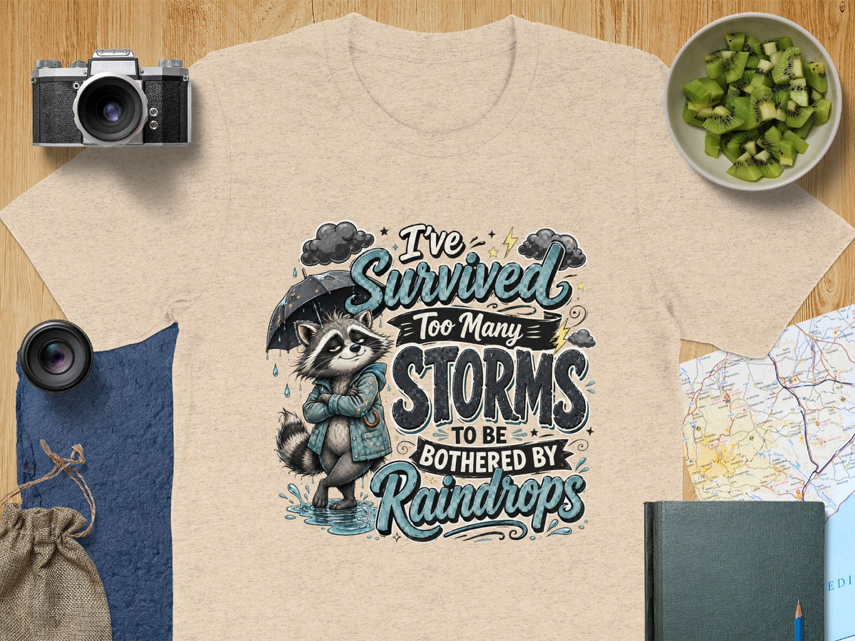 T-shirt graphic with raccoon holding umbrella, text "I've Survived Too Many STORMS TO BE BOTHERED BY Raindrops".