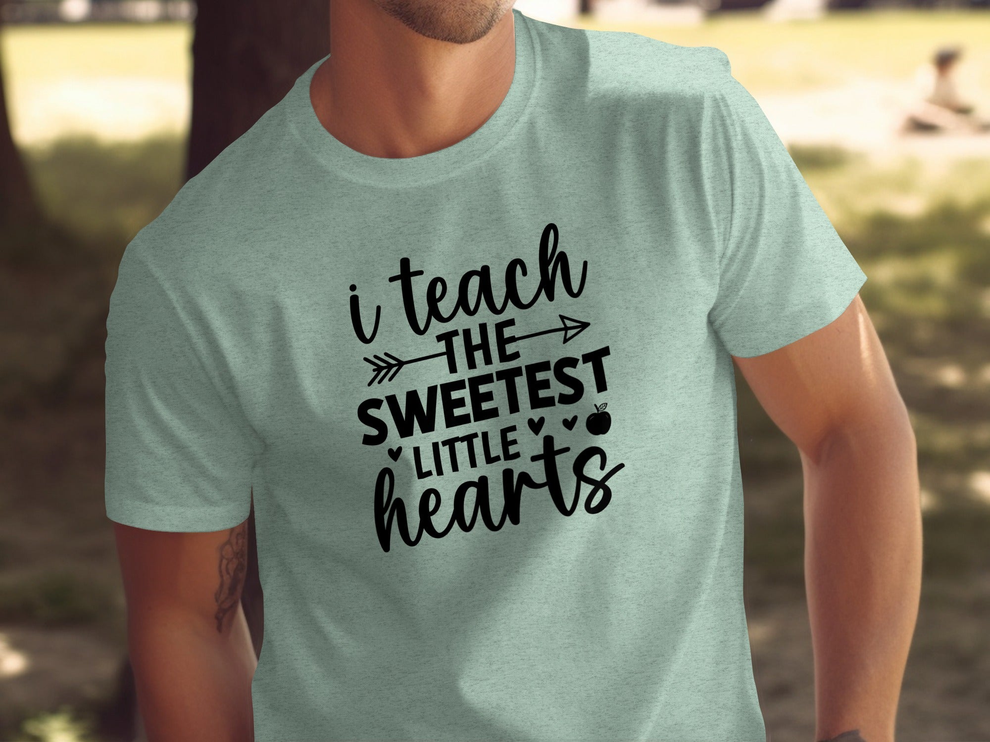 A man wears a light mint green t-shirt with black text reading "i teach THE SWEETEST LITTLE hearts".