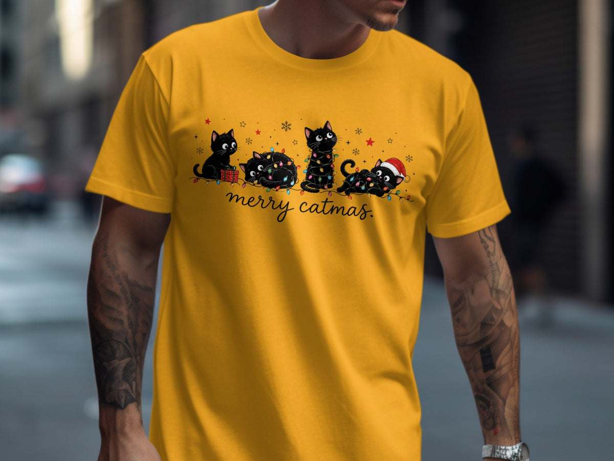 A person wears a yellow t-shirt featuring four cartoon black cats tangled in Christmas lights with the text merry catmas.