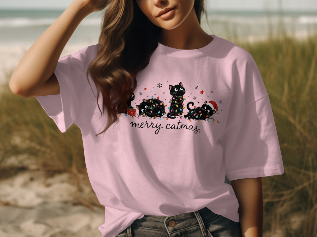 A person wearing a pink t-shirt with a graphic of black cats wrapped in Christmas lights and the text merry catmas.