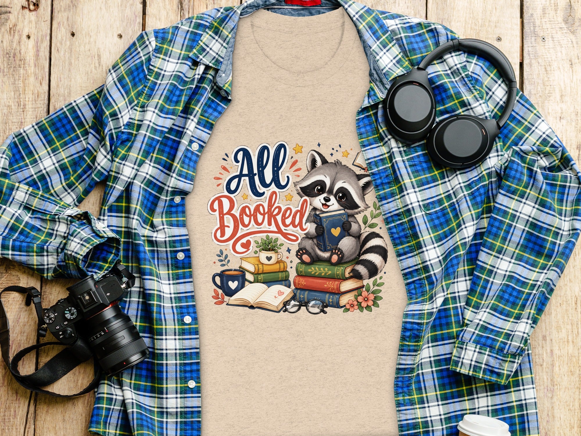 T-shirt with "All Booked" text and raccoon on books, laid flat with plaid shirt.