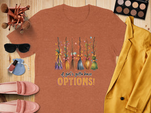 Orange t-shirt with colorful broomsticks graphic and text "A girl's gotta have OPTIONS!"
