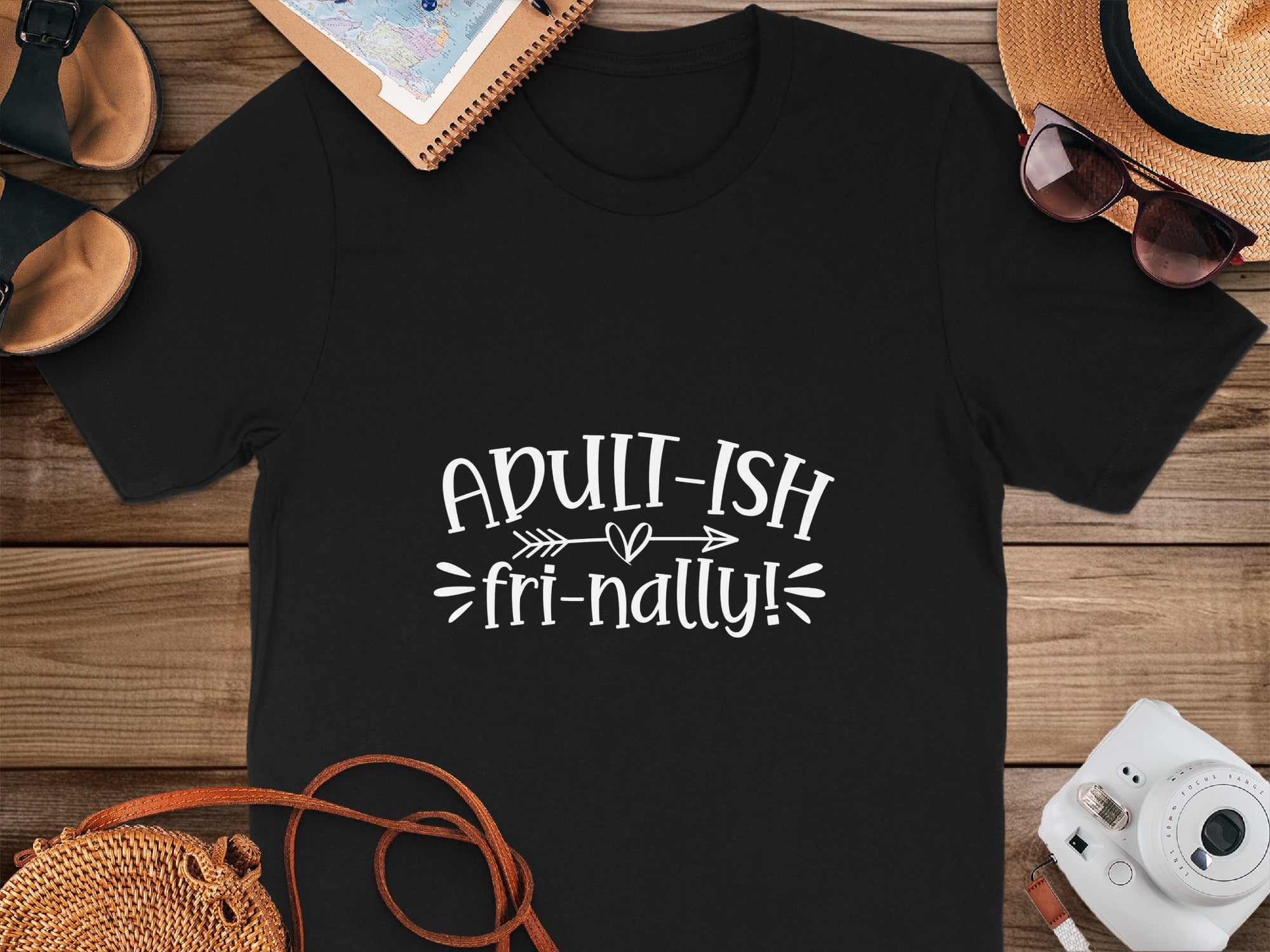 Black t-shirt with "ADULT-ISH fri-nally!" text, featuring travel accessories.