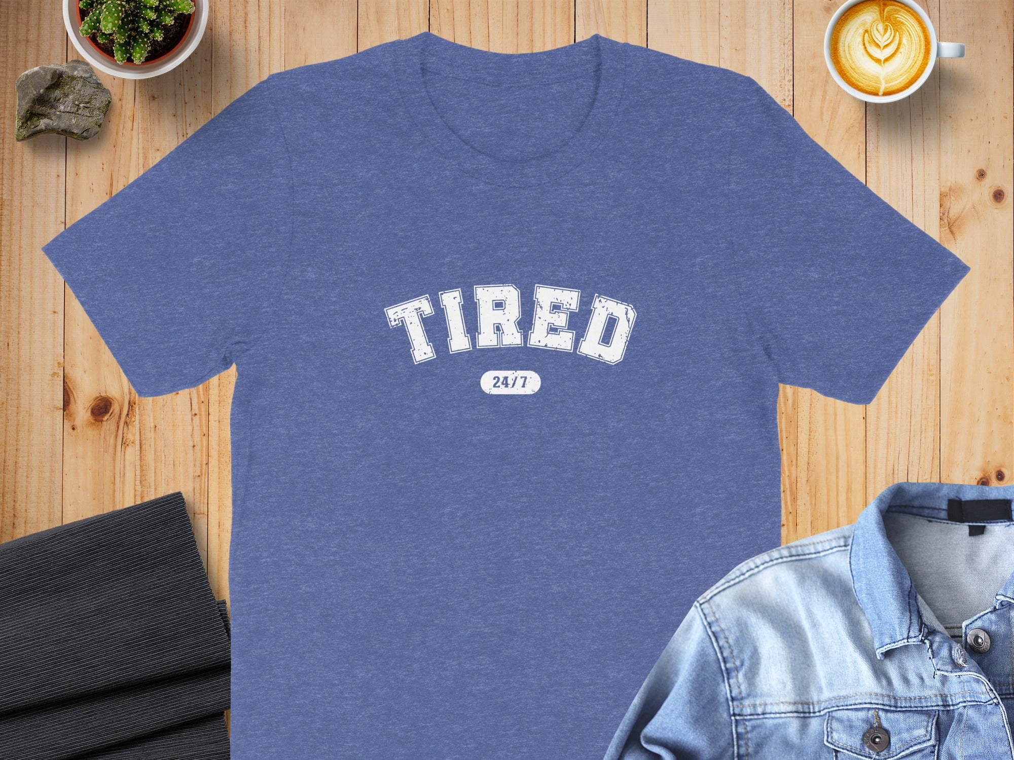 Blue t-shirt with "TIRED 24/7" graphic on a wooden surface.